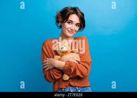 Ritratto fotografico di una graziosa ragazza che abbraccia un orsacchiotto elegante vestito marrone, isolato su sfondo blu Foto Stock