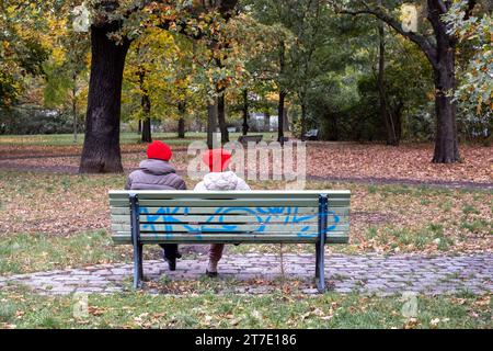 Zwei Frauen mit roten Mützen sitzen auf dem Berliner Falkplatz auf einer Parkbank. / Due donne in berretto rosso siedono su una panchina del parco sulla Falkplatz di Berlino. Herbst a Berlino *** due donne in berretto rosso siedono su una panchina del parco sulla Falkplatz di Berlino due donne in berretto rosso siedono su una panchina del parco sulla Falkplatz Herbst di Berlino snph20231105568.jpg Foto Stock