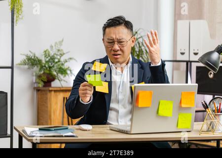 Uomo d'affari asiatico stanco e esausto che lavora su un computer portatile in ufficio con molte iscrizioni di attività con adesivi. L'uomo freelance impegnato nell'attività lavorativa sente una triste noia, un lavoro troppo impegnativo in multitasking. Attacco di panico, scadenza Foto Stock