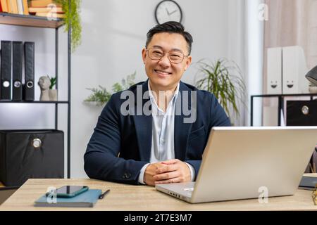 Ritratto di un felice uomo d'affari asiatico sorridente alla moderna scrivania dell'ufficio domestico che guarda la fotocamera. Cinese maschile in abito formale. Bell'uomo di mezza età lavora in remoto online su un notebook portatile Foto Stock