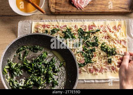 Il cuoco prepara mini torte per pizza, mette gli spinaci su sfoglia con formaggio e pancetta - vista dall'alto. Procedura di ricetta: 4 di 10 Foto Stock