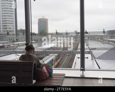 Giovane donna con panino, cappotto a scacchi e borse controlla e-mail sulla panchina di attesa alla stazione centrale di Utrecht con vista sui binari del treno Foto Stock
