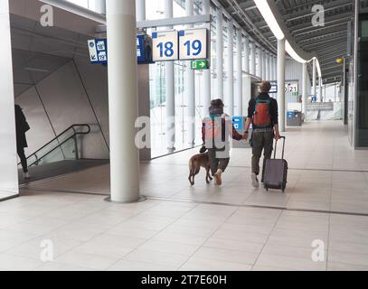 Giovane coppia viaggiatrice mano nella mano con pantaloni cargo, trolley, zaino rosso e cane in una passeggiata al guinzaglio attraverso l'edificio della stazione, Utrecht Centraal Foto Stock