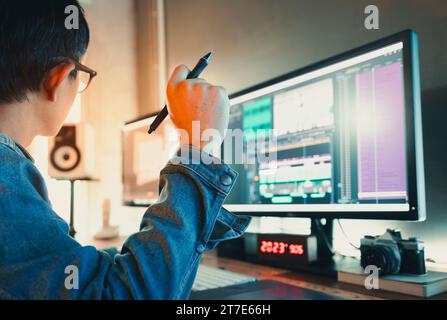 Video editor maschio asiatico che indossa una giacca in denim, lavora con filmati o video nel suo studio creativo. Foto Stock