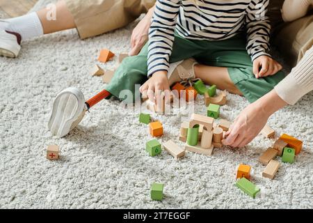 foto corta, ragazza con gamba protesica seduta sul tappeto e giocando con blocchi di legno vicino alla madre Foto Stock