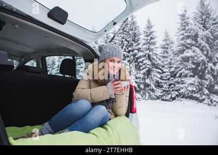 Donna con cappello di lana siede nel bagagliaio dell'auto e tiene una tazza di tè caldo in mano. Foto Stock