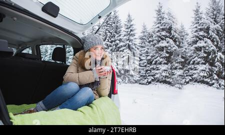 Donna con cappello di lana siede nel bagagliaio dell'auto e tiene una tazza di tè caldo in mano. Foto Stock