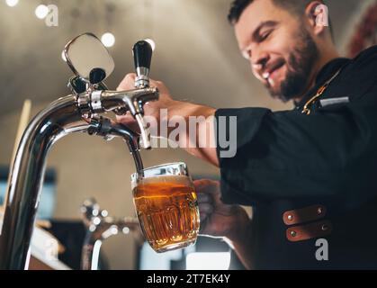 Assapora da vicino la birra lager fresca nella tazza di vetro. Barman barman barbarbato sorridente vestito in uniforme nera con un grembiule al bancone del bar. Persone di successo, Foto Stock