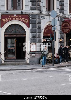 Copenaghen, Danimarca; 28 novembre 2022: Un ragazzo di origine asiatica si sta preparando ad attraversare una strada a Copenaghen e dietro di lui c'è un ristorante Foto Stock