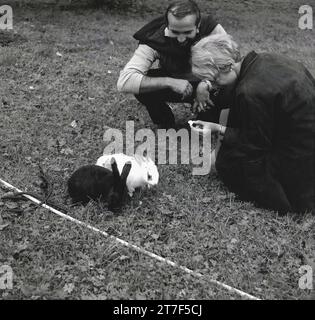 Anni '1960, un uomo e una donna inginocchiati su una sezione di erba con un pezzo di cibo per due conigli, uno nero e uno bianco, Inghilterra, Regno Unito. Foto Stock