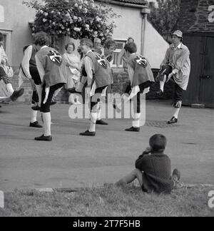 Anni '1970, storlcal, un ragazzino seduto a guardare uomini adulti e giovani uomini vestiti in costumi che fanno una danza morris, Inghilterra, Regno Unito. La danza Morris è una forma popolare di danza popolare inglese eseguita in festival e festività per celebrare l'arrivo dell'estate e il raccolto d'oro dell'autunno. Foto Stock