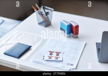 Concentrati su documenti cartacei, passaporto e foto del richiedente il visto sdraiato sulla scrivania con francobolli, matite e computer portatile Foto Stock
