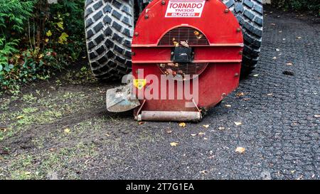 Primo piano di un soffiatore di foglie Tornado TM300 rosso attaccato a un veicolo di grandi dimensioni, che elimina le foglie autunnali dal percorso del parco, con un cartello di attenzione visibile. Foto Stock