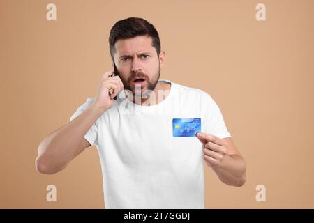 Uomo emotivo con carta di credito che parla su smartphone con sfondo beige. Attenzione - frode Foto Stock