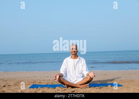 Senior indian Man seduto su un tappetino fitness in posizione di loto mentre meditava in spiaggia. Concetto di vita sana. Foto Stock
