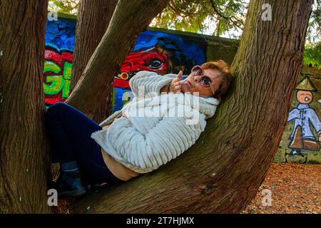 Giocosa donna anziana di oltre 60 anni, sdraiata sulla schiena nel tronco dell'albero di fronte al murale il giorno d'autunno a Hermitage Park, Helensburgh, Argyll e Bute, Scozia Foto Stock