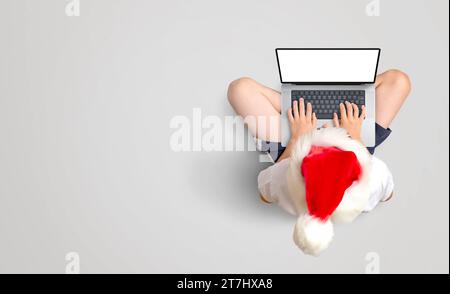 Il cappello di Natale ragazzo in rosso funziona su un computer portatile con schermo isolato per il mockup. Produttività festiva per terra. Copia spazio Foto Stock