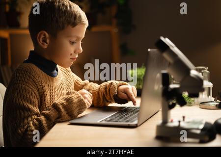 scolaro con un microscopio e un portatile sul posto di lavoro. Fare i compiti in biologia. Processo di insegnamento e ricerca scientifica Foto Stock