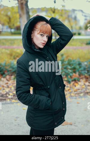 Donna rossa in cappotto nero con cappuccio che posa in autunno durante la passeggiata nella foresta Foto Stock