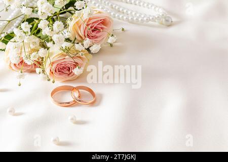 Organizzazione delicata del matrimonio con due anelli nuziali in oro su sfondo raso crema con perle e fiori. cartolina. invito. copertura Foto Stock