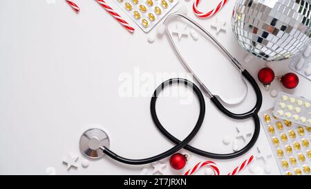 Banner medico di Natale.primo piano di stetoscopio, disco ball, lecca-lecca a righe, palline rosse, stelle e pillole su sfondo bianco, vista dall'alto, piatto, copia spa Foto Stock