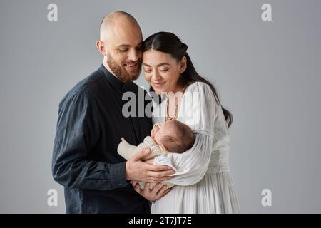 genitori moderni e gioiosi con abiti di classe sorridenti al loro neonato bambino su sfondo grigio, famiglia Foto Stock