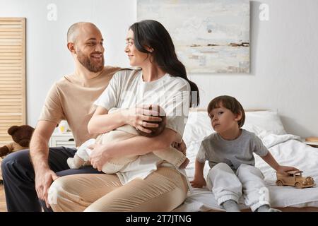 bambino con un giocattolo che guarda la macchina fotografica seduto accanto ai suoi genitori che tengono il fratello appena nato Foto Stock