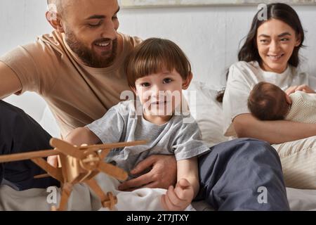 gioioso bambino in età prescolare seduto sul letto circondato dai suoi amorevoli genitori e dal suo fratello appena nato Foto Stock