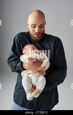 padre gioioso in un abbigliamento elegante che tiene il neonato addormentato su sfondo grigio, concetto di famiglia Foto Stock