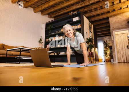 Una donna anziana attiva che fa yoga seguendo i passaggi del notebook. Donna anziana nel salotto di casa sua che fa un corso di yoga online. Foto Stock
