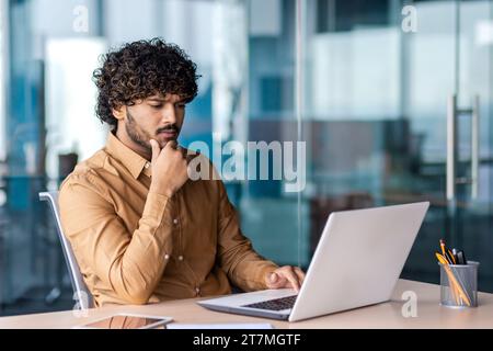 Uomo d'affari serio all'interno dell'ufficio sul posto di lavoro, si concentra e si concentra sulla risoluzione dell'attività corrente, il dipendente utilizza un notebook al lavoro, legge la documentazione online. Foto Stock