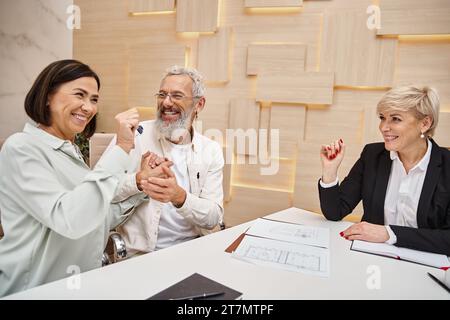 felice coppia sposata che tiene la chiave da una nuova casa vicino a un agente immobiliare sorridente nell'ufficio immobiliare Foto Stock