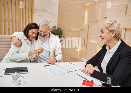 allegra coppia sposata che tiene la chiave da una nuova casa vicino a un agente immobiliare sorridente nell'ufficio immobiliare Foto Stock