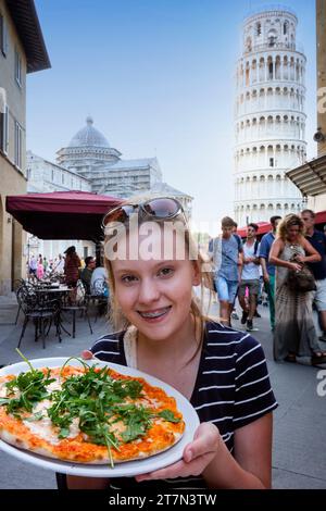 Adolescente con i capelli biondi, gli occhi noccioli e le bretelle dentali tiene una pizza fatta in casa in un caffè all'aperto con la Torre Pendente di Pisa nello sfondo Foto Stock