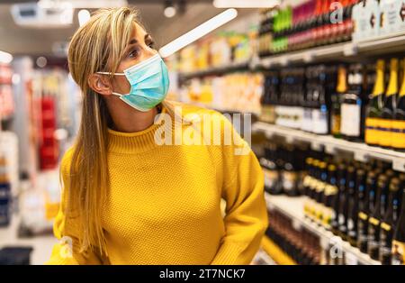 donna con maglione giallo e maschera che fa shopping nel corridoio del vino in negozio Foto Stock