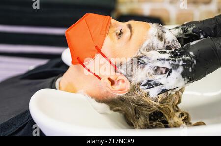 donne con maschera rossa ottenere il lavaggio dei capelli in un salone di bellezza. Parrucchiere che lava i capelli durante la pandemia di covid-19 Foto Stock