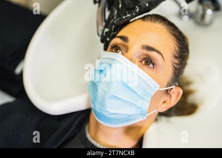 donna con maschera facciale si fa lavare i capelli in salone, professionale parrucchiere lavaggio capelli cliente con acqua e shampoo trattamento, durante co Foto Stock