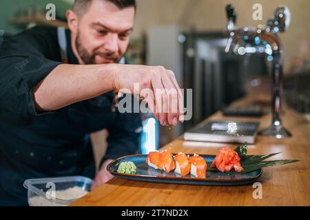 Cuoco barbuto vestito di uniforme nera che spruzza il piatto di sushi Nigirizushi nella cucina del ristorante nel seminterrato. Professione professionale, preparazione alimentare Foto Stock