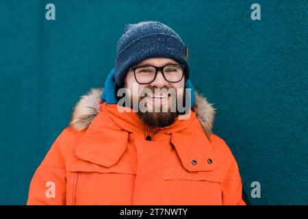 Ritratti bellissimi occhiali da vista giovane maschio barbato sorridente si erge su uno sfondo blu a parete in una giacca invernale arancione brillante con cappuccio con pelliccia Foto Stock