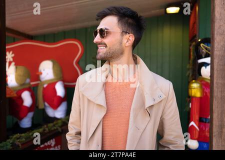 Uomo elegante con occhiali da sole sorride guardando lateralmente in piedi all'aperto. Uomo barbuto in piedi vicino alla decorazione natalizia all'esterno. Persone felici emozioni positive Foto Stock