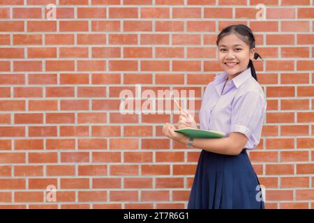 Ritratto di uno studente di scuola tailandese asiatico in uniforme sorriso con matita e libro tascabile per appunti Foto Stock