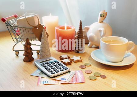 Risparmio di denaro per lo shopping natalizio, calcolatrice, salvadanaio, monete e banconote su un tavolo tra decorazioni natalizie, candele e carrello della spesa, copia Foto Stock