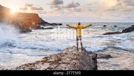 Persona in giacca gialla in piedi sulla roccia in riva al mare con le braccia aperte al tramonto Foto Stock