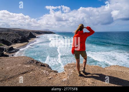Escursionista con giacca rossa che guarda le onde dell'oceano da una scogliera in una giornata di sole Foto Stock