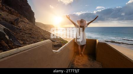 Donna con le braccia allungate alle scale della spiaggia, che si gode il tramonto sull'oceano a Playa de Cofete, Fuerteventura, Isole Canarie. Foto Stock