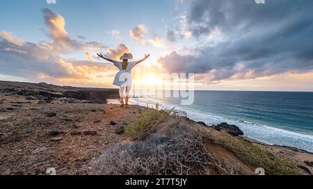 Donna in abito bianco e cappello con le braccia sollevate di fronte al mare al tramonto a Playa de Cofete, Fuerteventura, Isole Canarie. Foto Stock