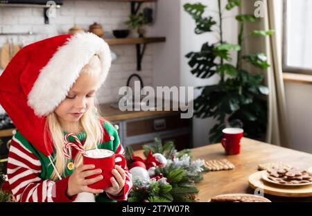 Bambino seduto sul bancone decorato a Natale. Bambino serio e guarda la tazza rossa con bevande e canne caramelle dentro Foto Stock