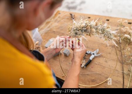 Primo piano delle mani di un fiorista che collega fiori secchi su un cerchio contro uno sfondo di legno Foto Stock