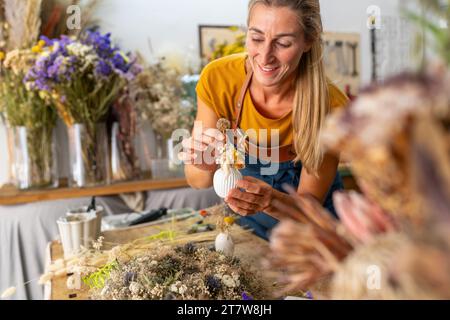 fiorista femminile che organizza un piccolo vaso bianco con fiori secchi in un laboratorio luminoso Foto Stock