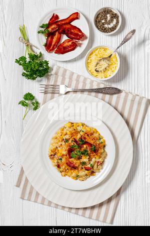 porzione di mac e formaggio con mais, pancetta ricoperta di pangrattato panko su piatto bianco su tavolo di legno bianco con ingredienti sullo sfondo, verticale Foto Stock